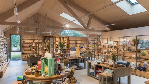 Retail shop interior with exposed timber beams, skylights, and wooden shelving displaying gifts, homewares, books, plants, and local products. Central display tables create an open, welcoming shopping space.